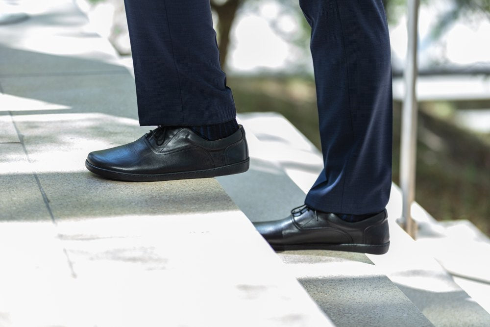 Une personne en pantalon de costume bleu foncé et chaussures SEQUENT noires de ZAQQ descend un escalier en pierre par une journée ensoleillée à l'extérieur. Le focus est sur les chaussures et la moitié inférieure des jambes, tandis que les arbres et la lumière du soleil forment un arrière-plan tacheté.