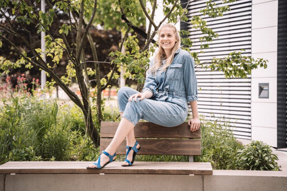 Une femme est assise sur un banc de jardin, vêtue d'une salopette en jean et de sandales pieds nus MIQA bleues avec une semelle en caoutchouc résistante de ZAQQ. Elle sourit dans la verdure, entourée d'arbres et de buissons.
