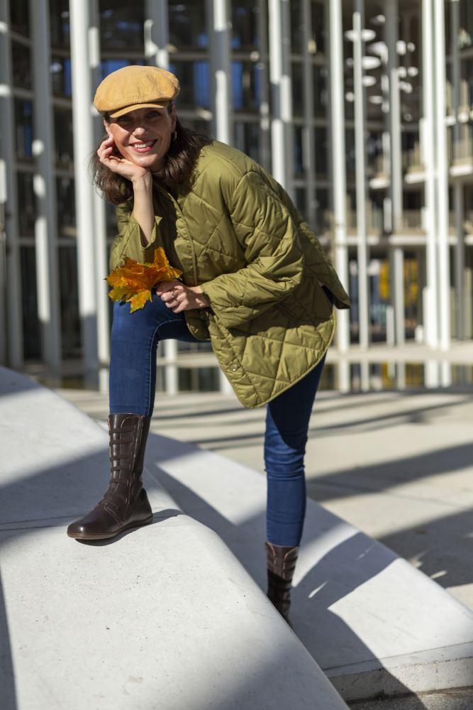 Eine Person mit beiger Mütze, grüner Steppjacke, Jeans und stylischem ATTRAQT Stretch Nappa Brown von ZAQQ steht im Freien, lehnt mit einem Knie auf einer weißen Struktur und hält gelbe Herbstblätter in den Händen. Sonnenlicht wirft Schatten von den umgebenden architektonischen Elementen und verleiht der Szene eine dynamische Note.